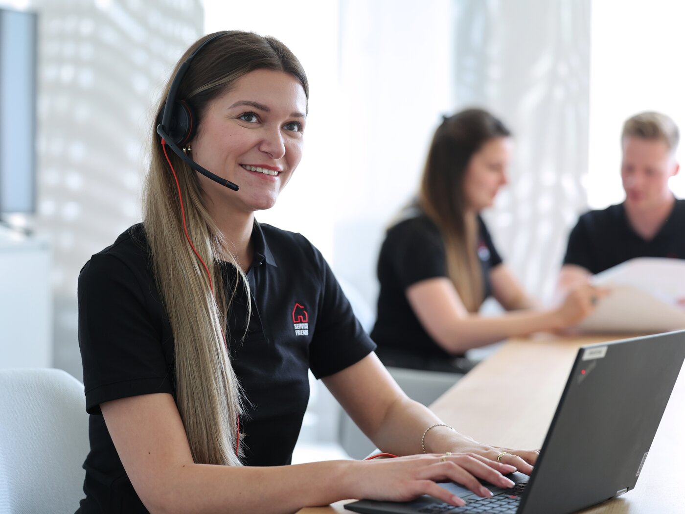 Eine Service Friends Mitarbeiterin, die ein Headset trägt, arbeitet an einem Laptop. Im Hintergrund sitzen zwei weitere Kollegen, die sich über Dokumente unterhalten.